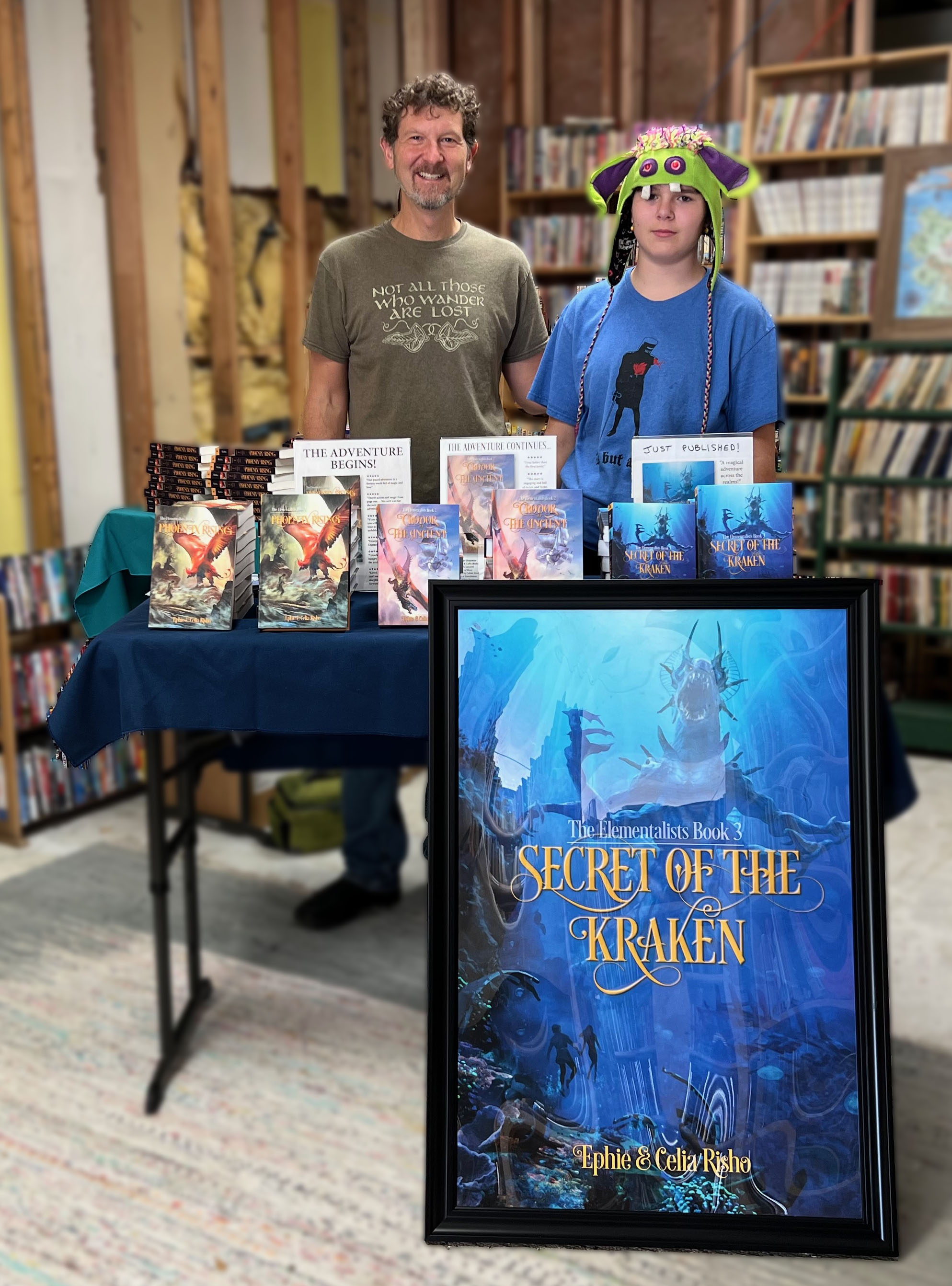 Ephie and Blue behind a book-signing table at Isle of Books, Blue wearing a knit monster hat, an oversize Secret of the Kraken poster on an easel